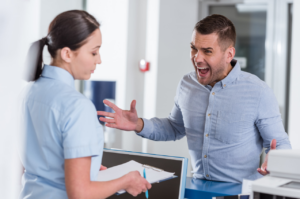 Frustrated patient yells at a practice receptionist, who looks down at her clipboard at the reception counter.