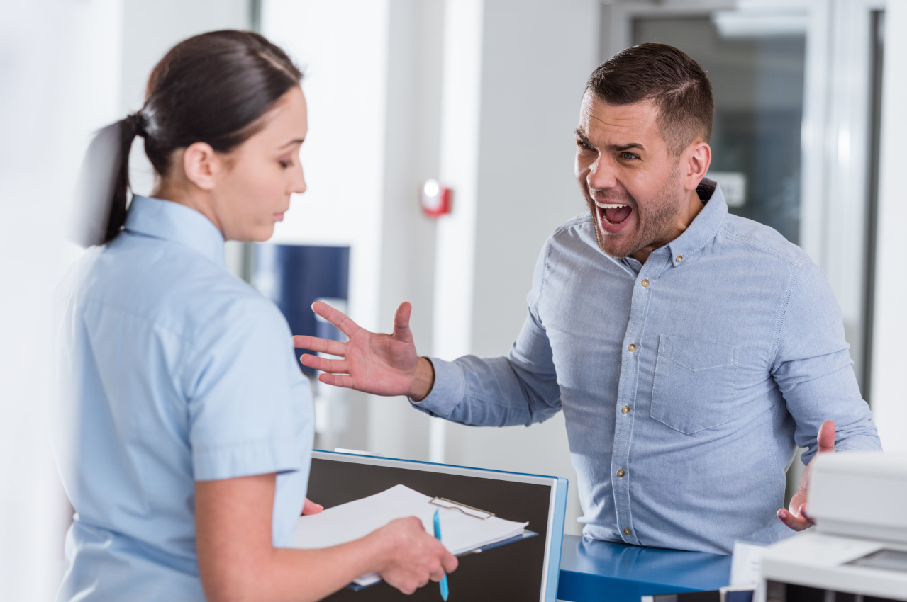 Frustrated patient yells at a practice receptionist, who looks down at her clipboard at the reception counter.