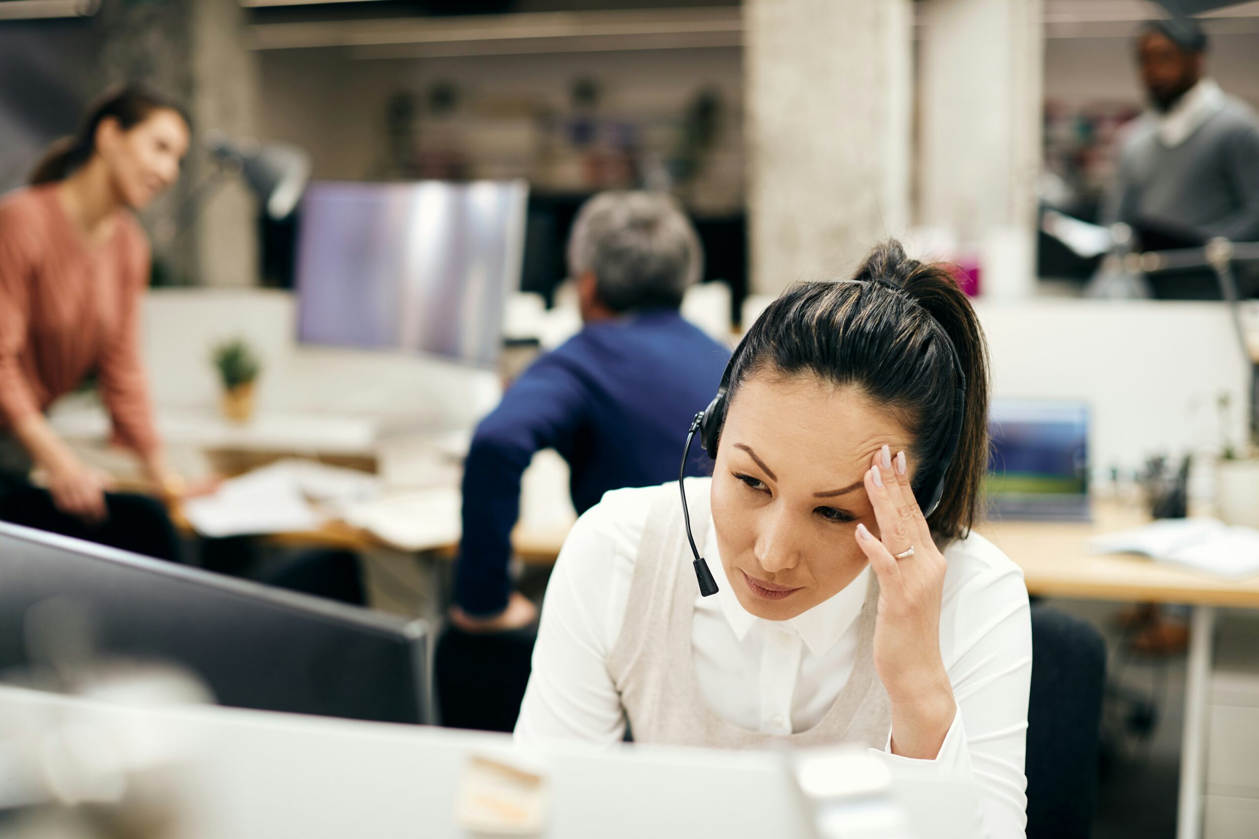 Receptionist wearing a headset holds her forehead in frustration while working at her computer in a busy clinic.